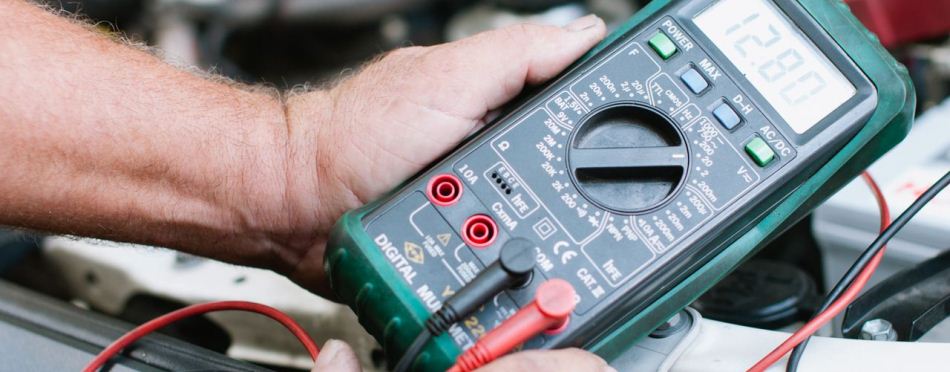 Hands holding a digital multimeter over an engine bay